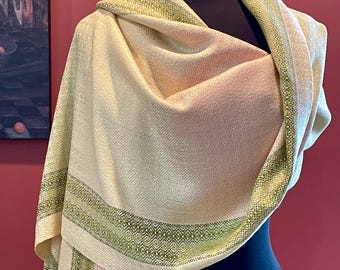 May include: A woven shawl featuring a cream-colored base with a subtle geometric diamond pattern. The fabric is accented by horizontal bands of variegated green and yellow-green threads, creating a textured border along the edges of the wrap.