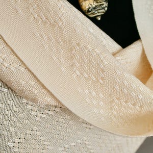 May include: A cream-coloured woven shawl with a delicate pattern. The shawl is folded in half and has a soft, drapey texture.