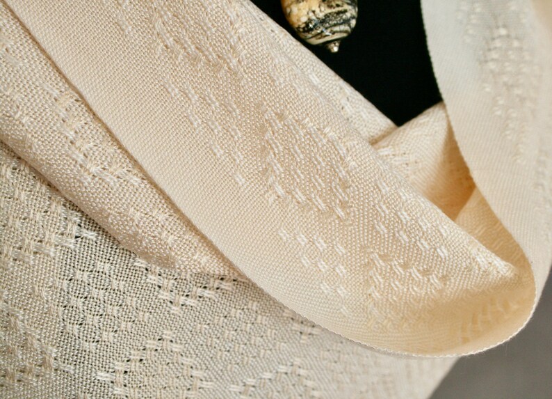 May include: A cream-coloured woven shawl with a delicate pattern. The shawl is folded in half and has a soft, drapey texture.