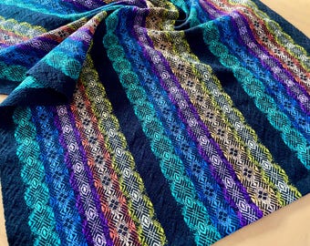 May include: A colorful woven textile with a striped pattern. The design features diagonal stripes in shades of blue, green, purple, orange, and yellow, set against a dark blue background. The fabric has a textured appearance.