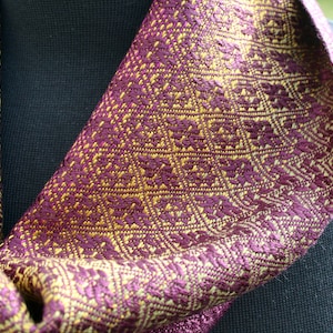 Pode incluir: Um cachecol roxo e dourado com um padr&atilde;o geom&eacute;trico. O cachecol est&aacute; dobrado e drapeado sobre uma superf&iacute;cie preta.