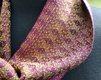May include: A purple and gold patterned scarf with a geometric design. The scarf is folded and draped over a black surface.
