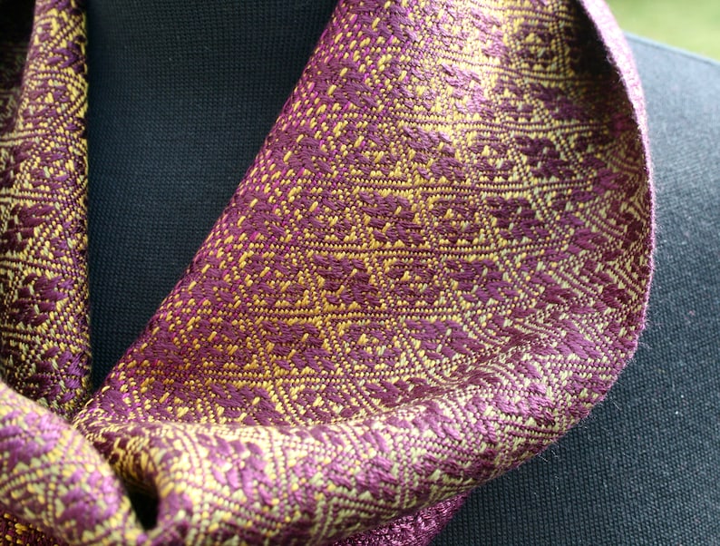 Pode incluir: Um cachecol roxo e dourado com um padr&atilde;o geom&eacute;trico. O cachecol est&aacute; dobrado e drapeado sobre uma superf&iacute;cie preta.