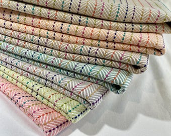 May include: A stack of folded fabric featuring a herringbone weave pattern in various pastel colors including pink, yellow, green, blue, and peach. Each layer is accented with vertical rows of small, multicolored embroidered stitches.