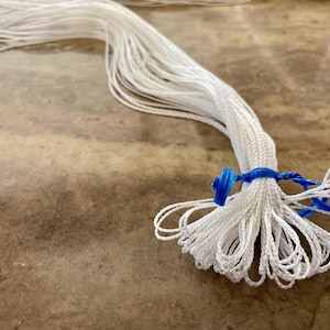 May include: Close-up of a bundle of white cord, secured with blue string and a blue plastic component. The cord is fanned out, with loops at the end. The background is a mottled brown surface.
