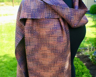 May include: A woven scarf with a geometric pattern in shades of brown, blue, and gold. The scarf has a fringe on the bottom edge.