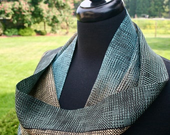 May include: A woven infinity scarf in shades of black, teal, and tan. The scarf is displayed on a black mannequin and features a textured, geometric pattern. The color palette and design suggest a handmade accessory.