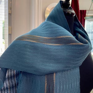 May include: A teal woven scarf featuring a subtle chevron texture and horizontal stripes in dark blue and metallic gold. The fabric displays a consistent weave pattern throughout, with the stripes running parallel along the length of the material.