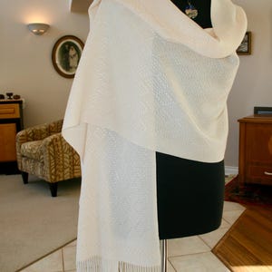 May include: A white shawl with a delicate pattern and fringe. The shawl is draped over a black mannequin.
