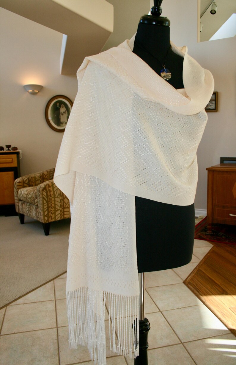 May include: A white shawl with a delicate pattern and fringe. The shawl is draped over a black mannequin.