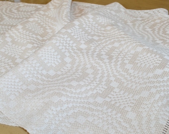 May include: A folded beige and white patterned textile. The fabric has a woven design with a repeating floral or geometric pattern. The textile is draped, showing the texture and folds of the material.