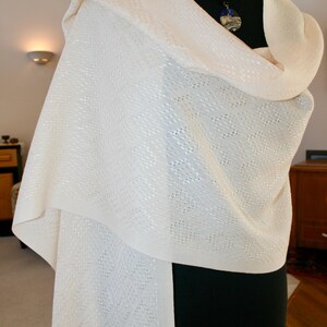 May include: A white knitted shawl with a delicate pattern. The shawl is draped over a black mannequin and has a blue and white pendant necklace.