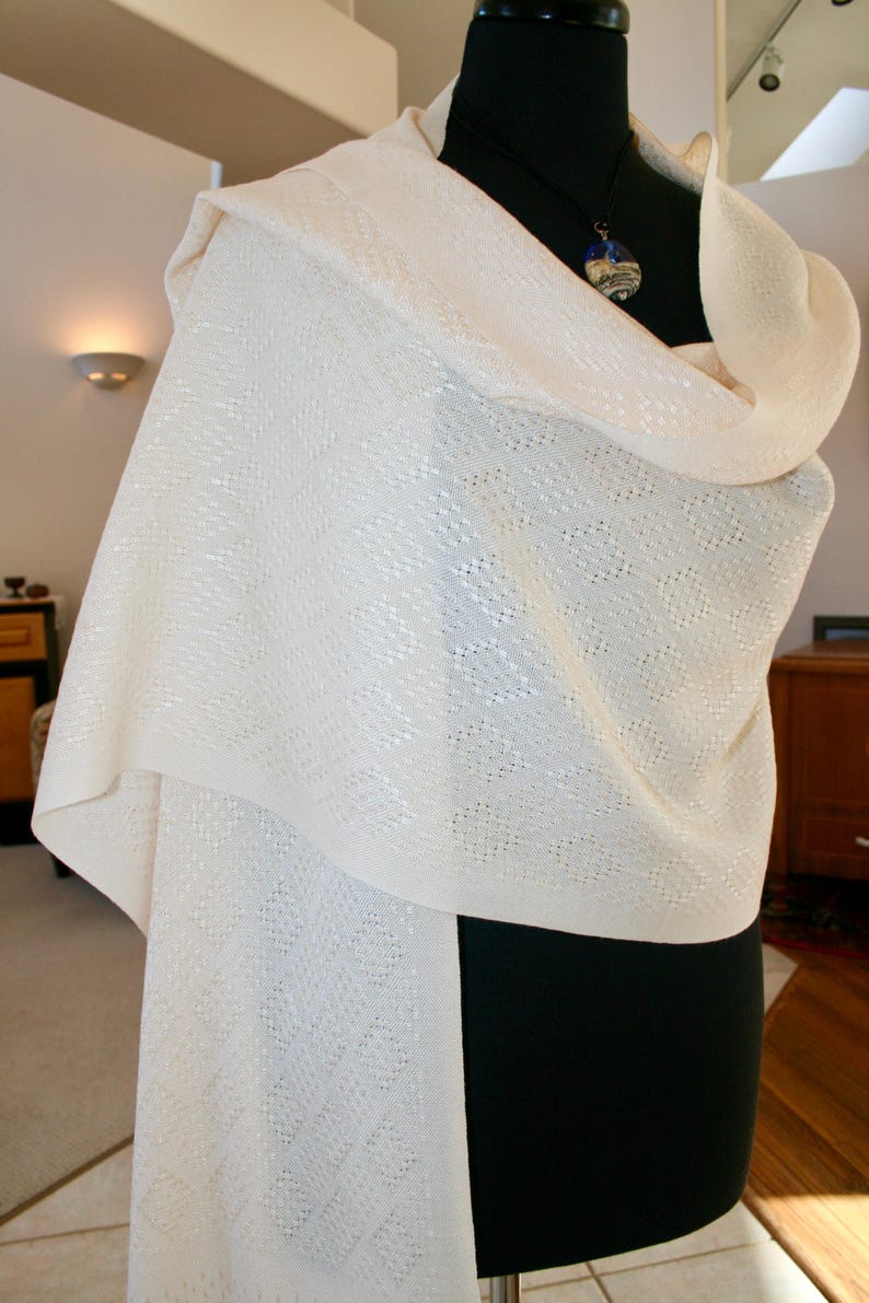 May include: A white knitted shawl with a delicate pattern. The shawl is draped over a black mannequin and has a blue and white pendant necklace.