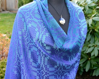 May include: A blue and green patterned shawl with a geometric design. The shawl is draped over a black mannequin and has a silver pendant necklace with a blue and white glass bead.