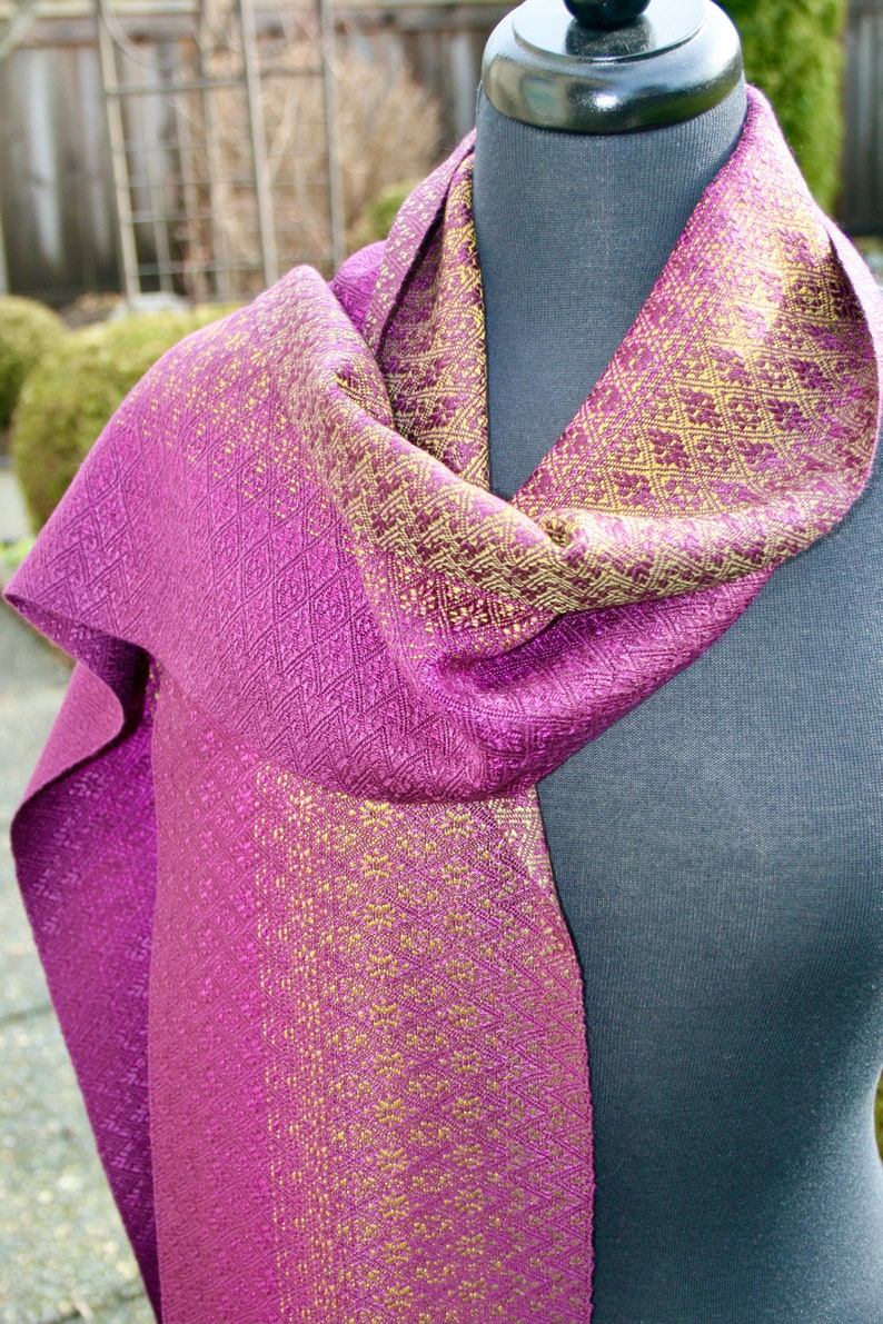 Pode incluir: Um cachecol de seda roxo e dourado com um padr&atilde;o geom&eacute;trico. O cachecol est&aacute; drapeado sobre um manequim.