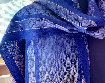 May include: A blue scarf with a leaf pattern and a striped design. The scarf is draped over a mannequin. The scarf has a detailed pattern in a lighter color, creating a contrast against the blue fabric.