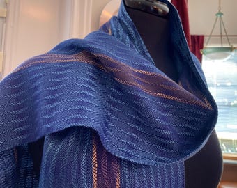 May include: A woven scarf featuring a deep blue and light blue chevron pattern with vertical copper-colored stripes. The fabric has a textured appearance with a visible weave structure, draped over a dark form to display the intricate textile design.