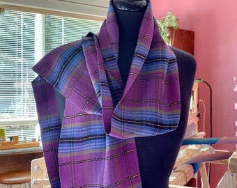 May include: A purple and blue plaid scarf is draped over a mannequin. The scarf has a woven design with stripes of purple, blue, yellow, and black. The scarf is made of a soft material and is a fashionable accessory.