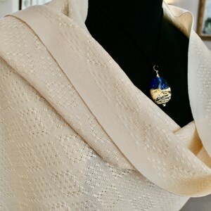 May include: A cream-coloured shawl with a delicate woven pattern. A silver chain with a blue and white glass pendant hangs from the shawl.