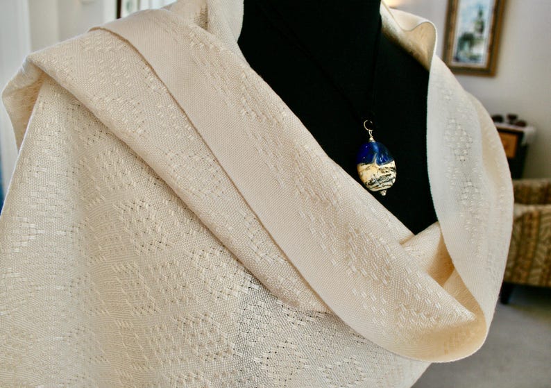 May include: A cream-coloured shawl with a delicate woven pattern. A silver chain with a blue and white glass pendant hangs from the shawl.
