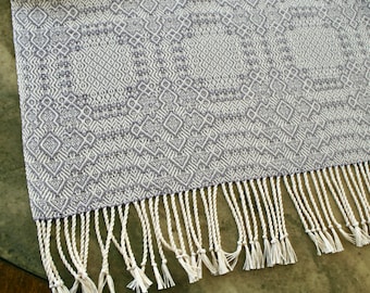 May include: A white and gray woven table runner with a geometric pattern and a fringe border.