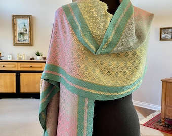 May include: A woven shawl in pastel colors of pink, green, and yellow, displayed on a black mannequin. The shawl features a diamond pattern and striped borders. The ends of the shawl have tassels.