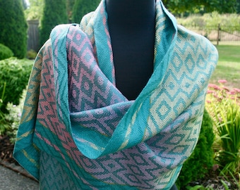 May include: A teal, pink, and gold woven shawl with a geometric diamond pattern. The scarf is draped over a black mannequin, showcasing the intricate design and color variations. The soft fabric appears to be lightweight.