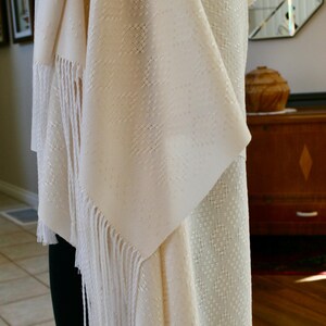 May include: A white shawl with a delicate woven pattern and fringe. The shawl is draped over a mannequin and has a soft, flowing texture.