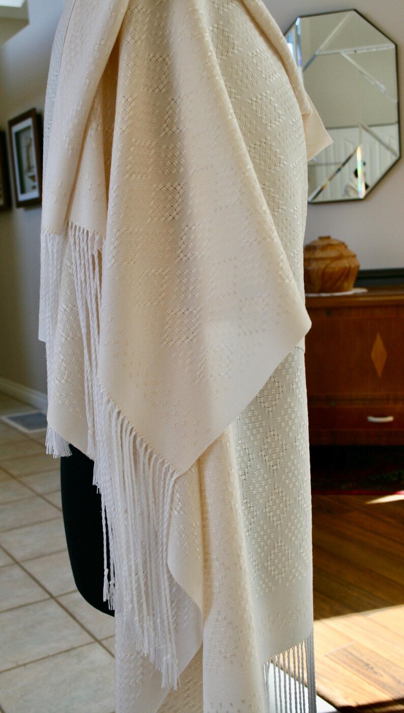 May include: A white shawl with a delicate woven pattern and fringe. The shawl is draped over a mannequin and has a soft, flowing texture.