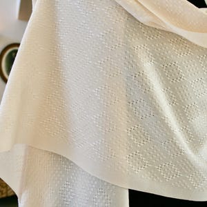 May include: A white knitted shawl with a delicate diamond pattern. The shawl is soft and lightweight, making it perfect for layering or wearing on its own.