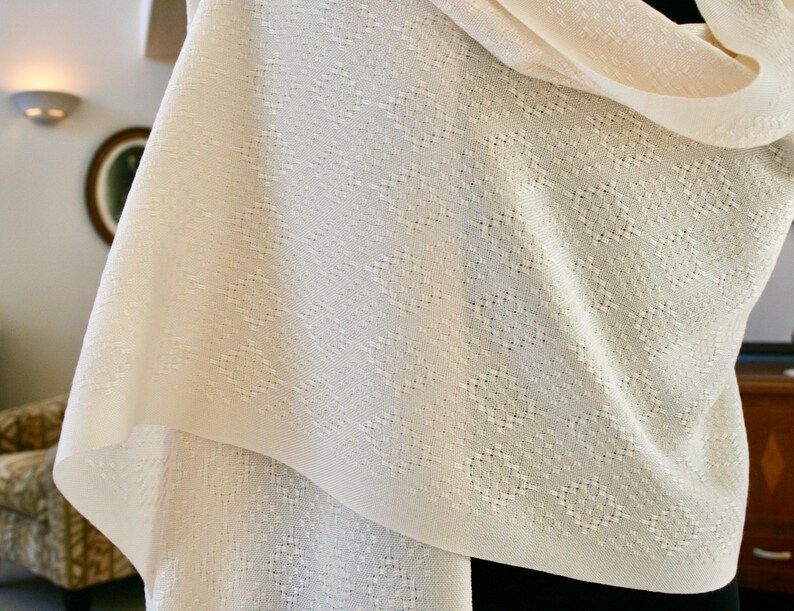 May include: A white knitted shawl with a delicate diamond pattern. The shawl is soft and lightweight, making it perfect for layering or wearing on its own.