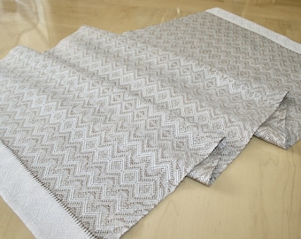 May include: A folded, neutral-toned table runner with a repeating diamond pattern. The runner has a textured appearance and a white border on each end. The fabric is a light beige color with a geometric design.