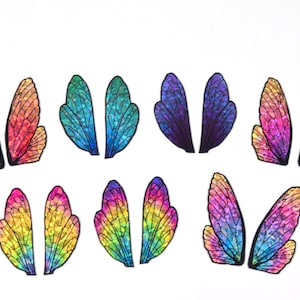 Puede incluir: Conjunto de ocho alas de hada arcoíris iridiscentes con contornos negros. Las alas están dispuestas en dos filas de cuatro, con cada fila presentando un degradado de color diferente.