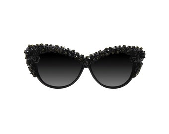 May include: Black cat-eye sunglasses with a decorative floral design along the top frame. The sunglasses have dark lenses and are adorned with small, dark green embellishments. The frame is black and the overall style is elegant.