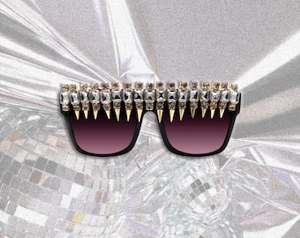 Puede incluir: Gafas de sol negras con un borde superior decorativo con cristales rectangulares y cuadrados transparentes, alternando con puntas doradas. Las lentes tienen un degradado de morado oscuro a claro. El fondo es un material plateado brillante.