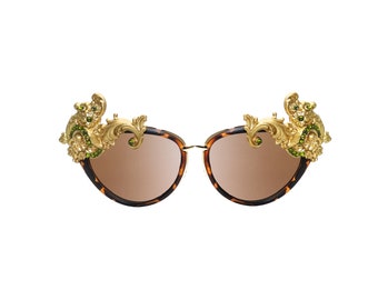 May include: Cat-eye sunglasses with a tortoiseshell pattern frame and brown lenses. The sunglasses feature ornate gold-colored embellishments with green gemstones at the top corners, adding a touch of luxury and style.