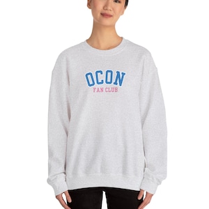 Puede incluir: Una sudadera gris con un gráfico azul y rosa que dice "OCON FAN CLUB".