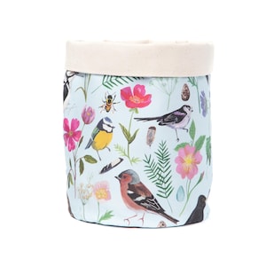 May include: A cylindrical fabric storage basket with a pale blue background. The basket is decorated with colourful illustrations of birds, flowers, and insects. The top edge is a natural canvas colour.