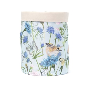 May include: A cylindrical storage basket with a light blue background, featuring a floral and wildlife design. The design includes blue flowers, butterflies, and a small bird. The top edge of the basket is a natural beige colour.