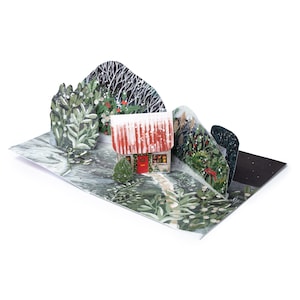 May include: A pop-up Christmas card depicting a snow-covered cottage with a red door and roof. The card shows a winter forest scene with trees and a path leading to the cottage. The card is open and displayed on a white surface.