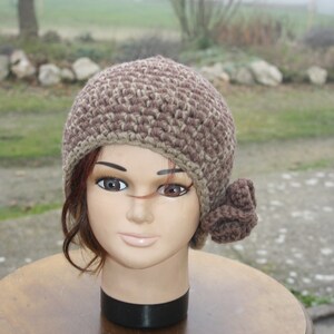Peut inclure: Un chapeau au crochet marron et beige avec une fleur décorative. Le chapeau est présenté sur une tête de mannequin avec des cheveux bruns, posé sur une table en bois. L'arrière-plan est une scène extérieure floue.