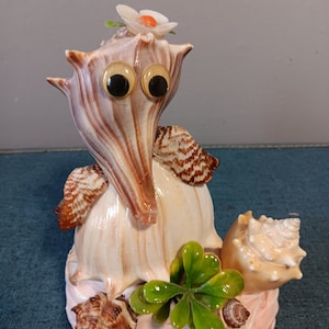May include: A whimsical seashell sculpture with googly eyes, a white flower, and green leaves. The sculpture is made of various seashells and is sitting on a pink base.