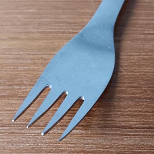 May include: A stainless steel fork with four tines, angled slightly to the left. The fork is set against a wooden background, with the tines pointing towards the bottom left corner. The fork's handle is smooth and rounded.
