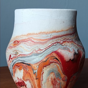 May include: A handcrafted ceramic vase with a unique, abstract design. The upper portion is white, transitioning to a vibrant pattern of orange, red, gray, and black swirls. The vase has a rounded body and a slightly flared rim.