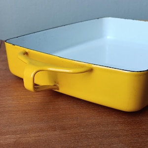 May include: A yellow enamel baking dish with a white interior and a single handle. The dish is sitting on a wooden surface.