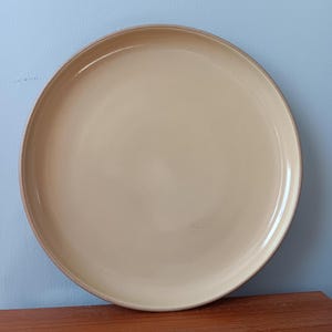 May include: A large, round, beige ceramic plate with a slightly raised rim. The plate has a smooth, glossy finish and a simple, minimalist design. The plate is suitable for serving meals.