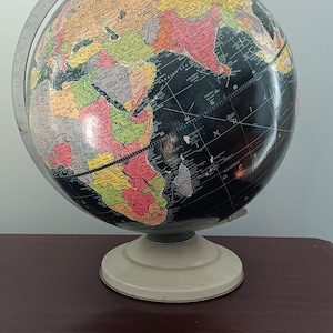 May include: A vintage globe with a black ocean and colorful continents. The globe is mounted on a cream-colored base and has a silver meridian ring. The globe is displayed on a dark wood surface.