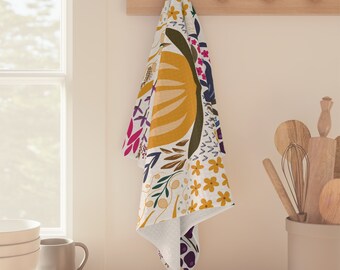 Könnte beinhalten: Ein weißes Küchentuch mit farbenfrohem Blumenmuster hängt an einer Holzleiste. Das Design umfasst große gelbe, grüne und braune Formen sowie kleinere Blüten und Blätter in Rosa, Blau und Gelb. Das Tuch hängt in einer Küche.