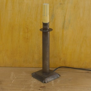 May include: A black metal candlestick with a square base and a tall, cylindrical stem. A white candle sits in the top of the candlestick.