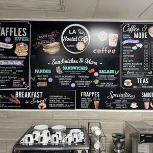 May include: A large black menu board for LA Social Cafe featuring white text and colorful illustrations of waffles, sandwiches, coffee, and smoothies. The menu lists various breakfast items, paninis, salads, frappes, and teas with pricing details.